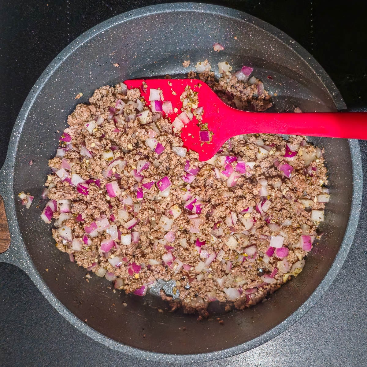 A medium saucepan with ground bison, onion, garlic, and cumin with a red spatula.