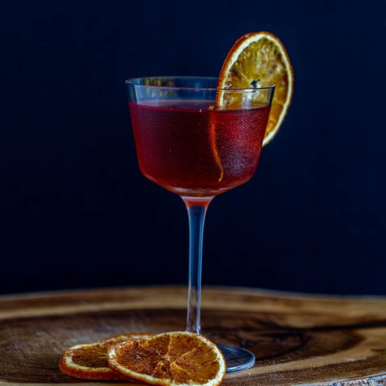 A single tepache cocktail with bourbon in a Nick and Nora glass with dehydrated orange wheel on a wood tray.