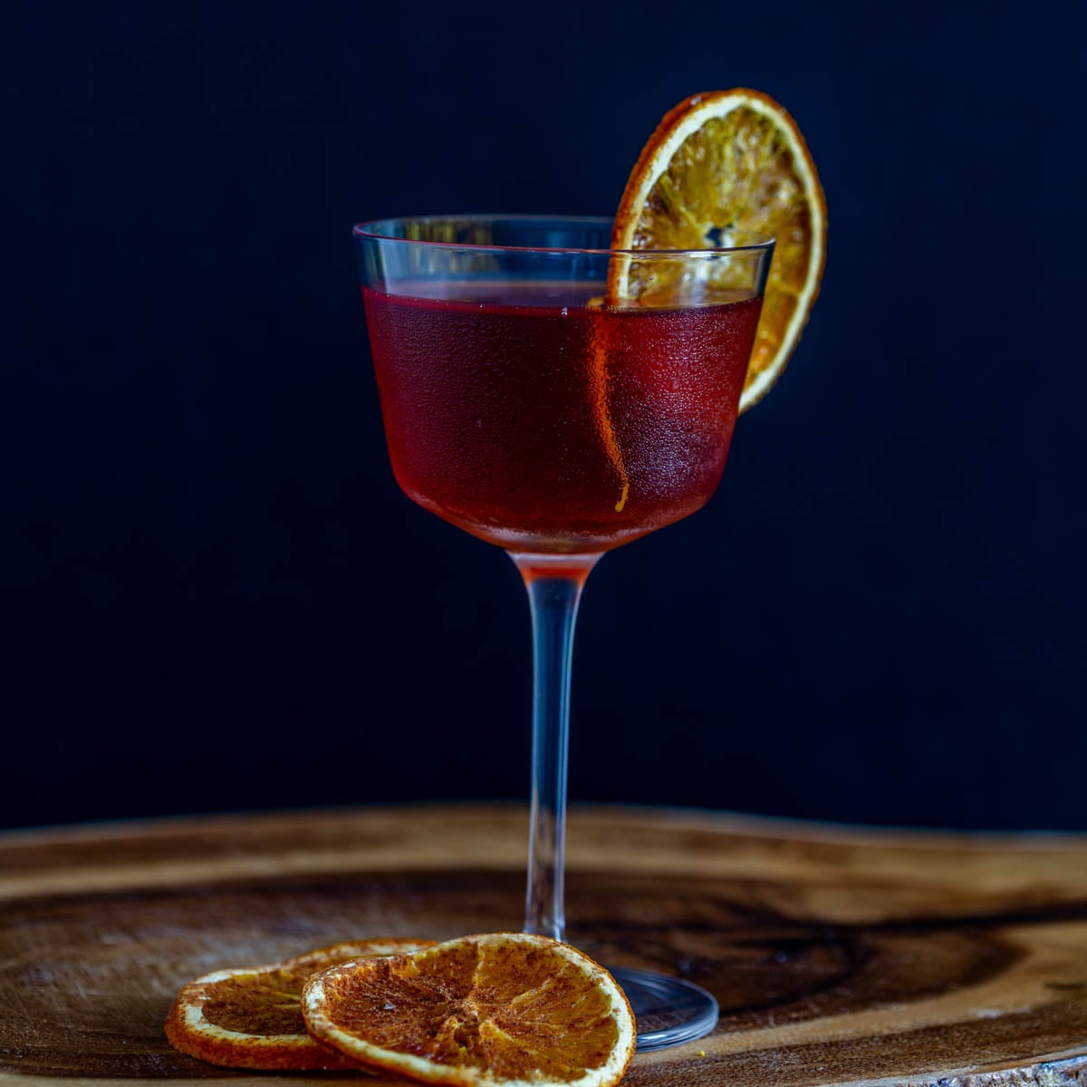 A single tepache cocktail with bourbon in a Nick and Nora glass with dehydrated orange wheel on a wood tray.