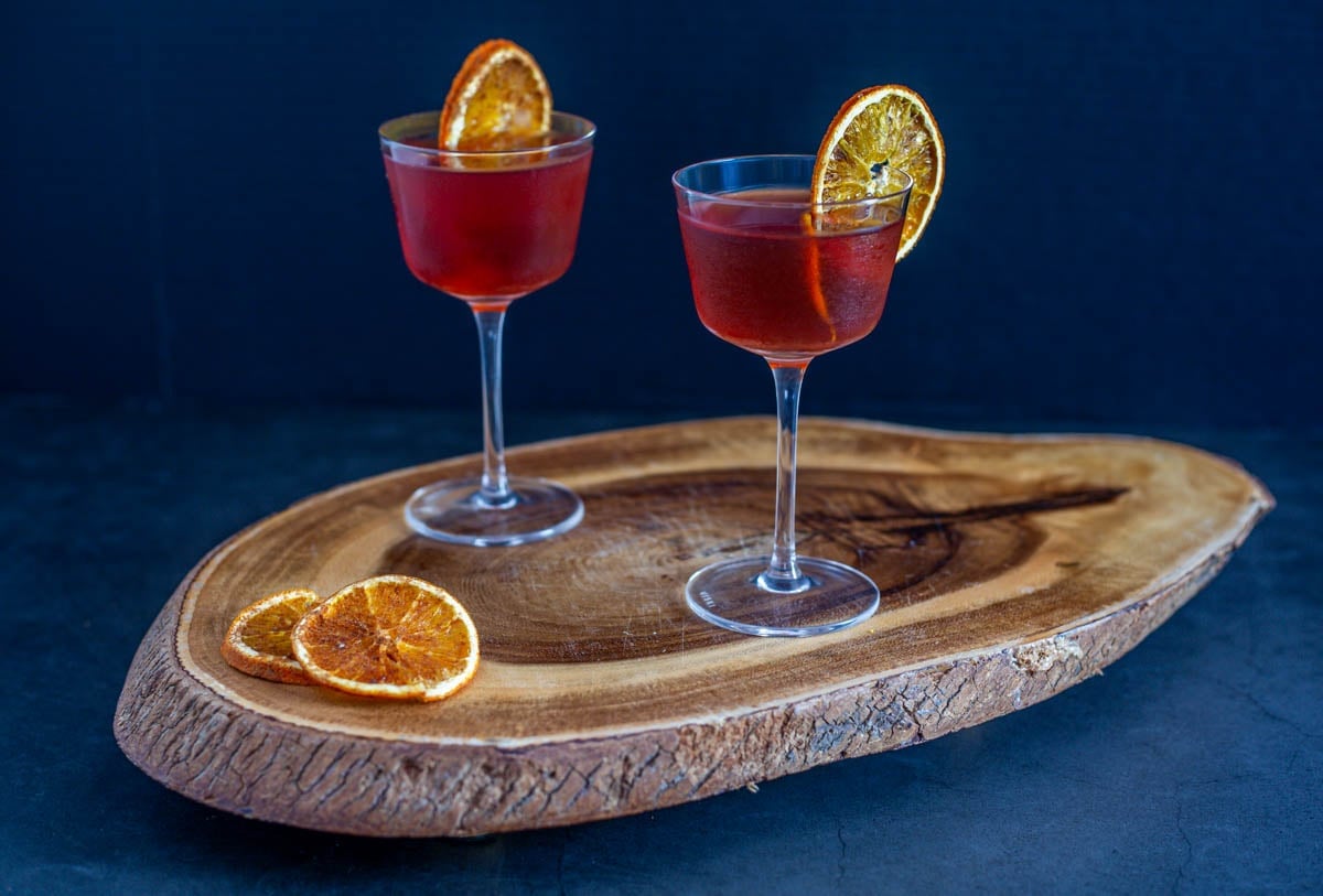 2 tepache cocktails with bourbon on a live edge wood tray with dehydrated orange wheels.