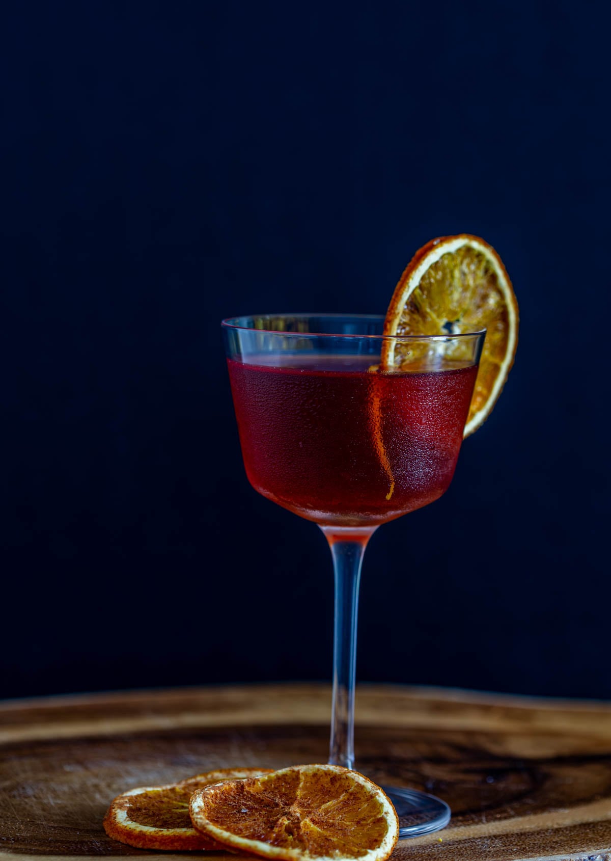 A single tepache cocktail with bourbon in a Nick and Nora glass with dehydrated orange wheel on a wood tray.