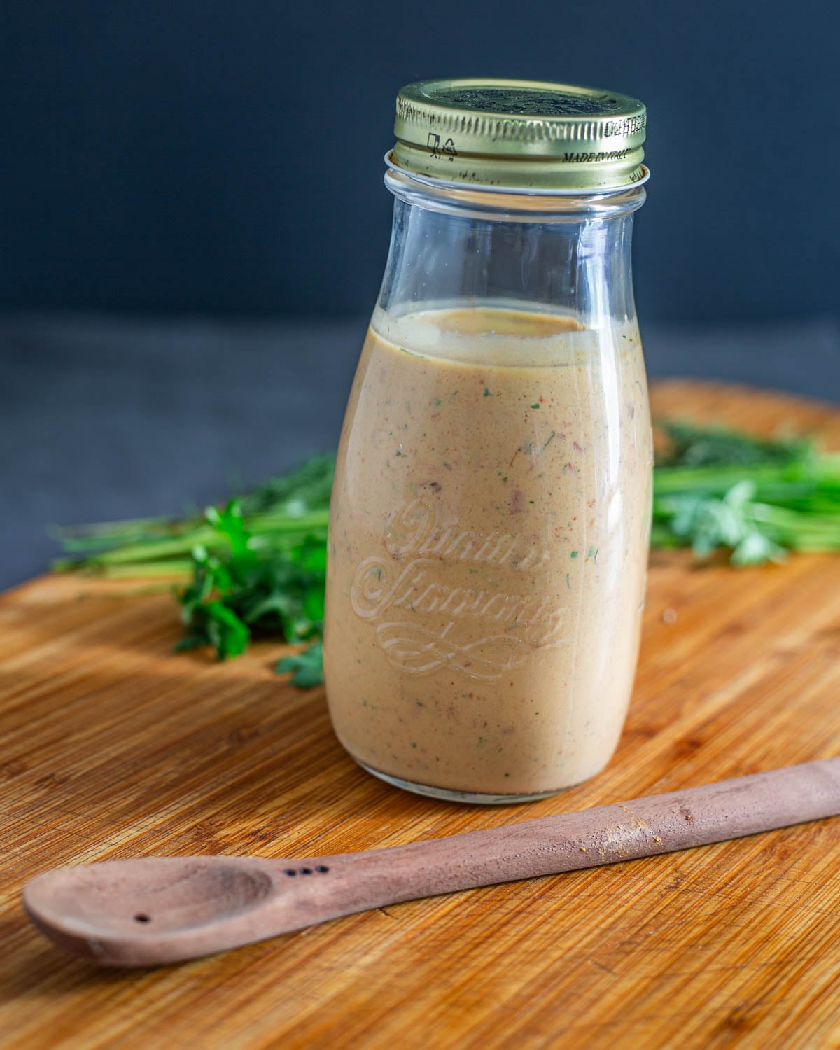 The completed bottle of chipotle ranch dressing in a glass bottle with herbs, wood spoon, and wood cutting board.