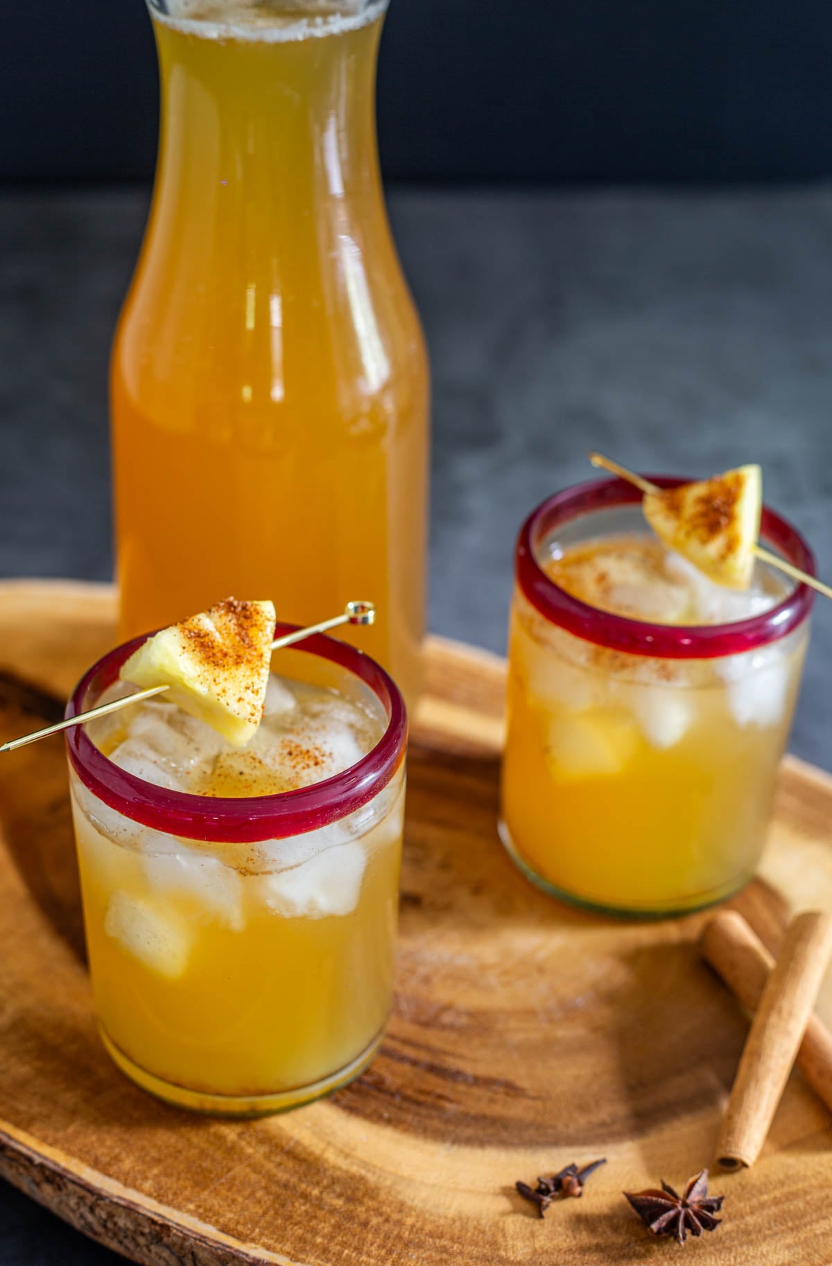 A carafe of homemade tepache with 2 red-rimmed glasses with ice, tepache, and pineapple cube.