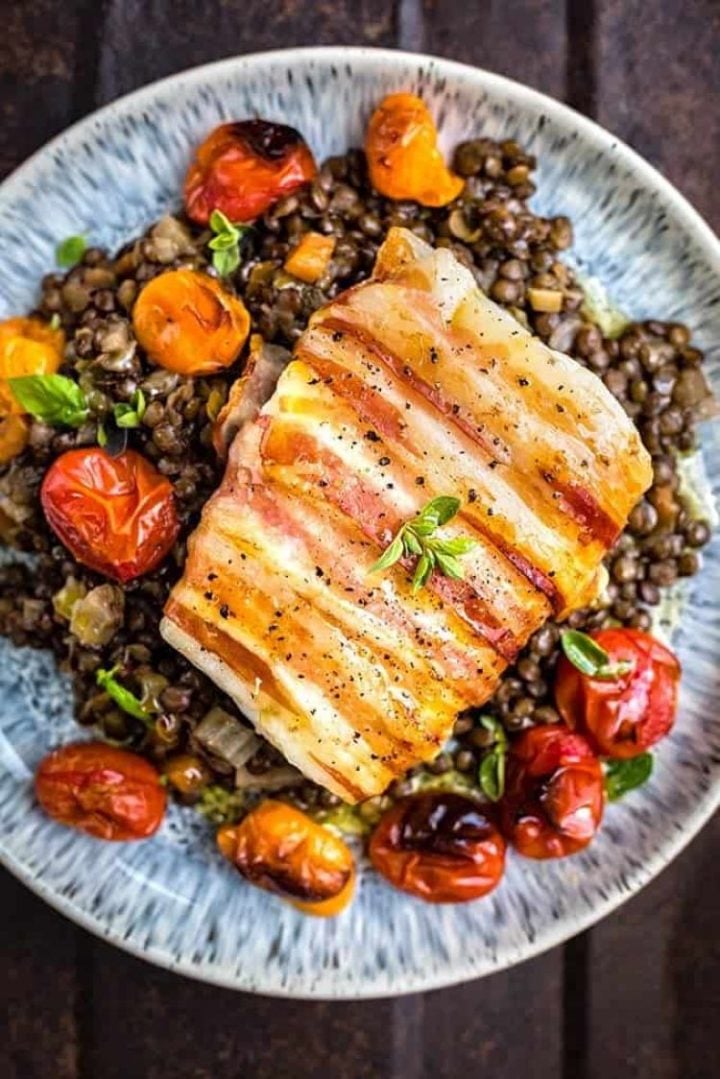 Pancetta wrapped cod with lentils and tomatoes on a stoneware plate.