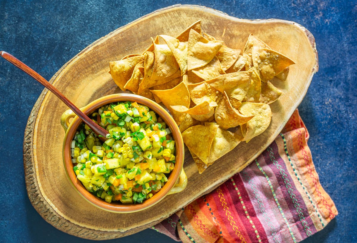 A live edge wood cutting board with a bowl of grilled corn and pineapple salsa and air-fryer corn tortilla chips.