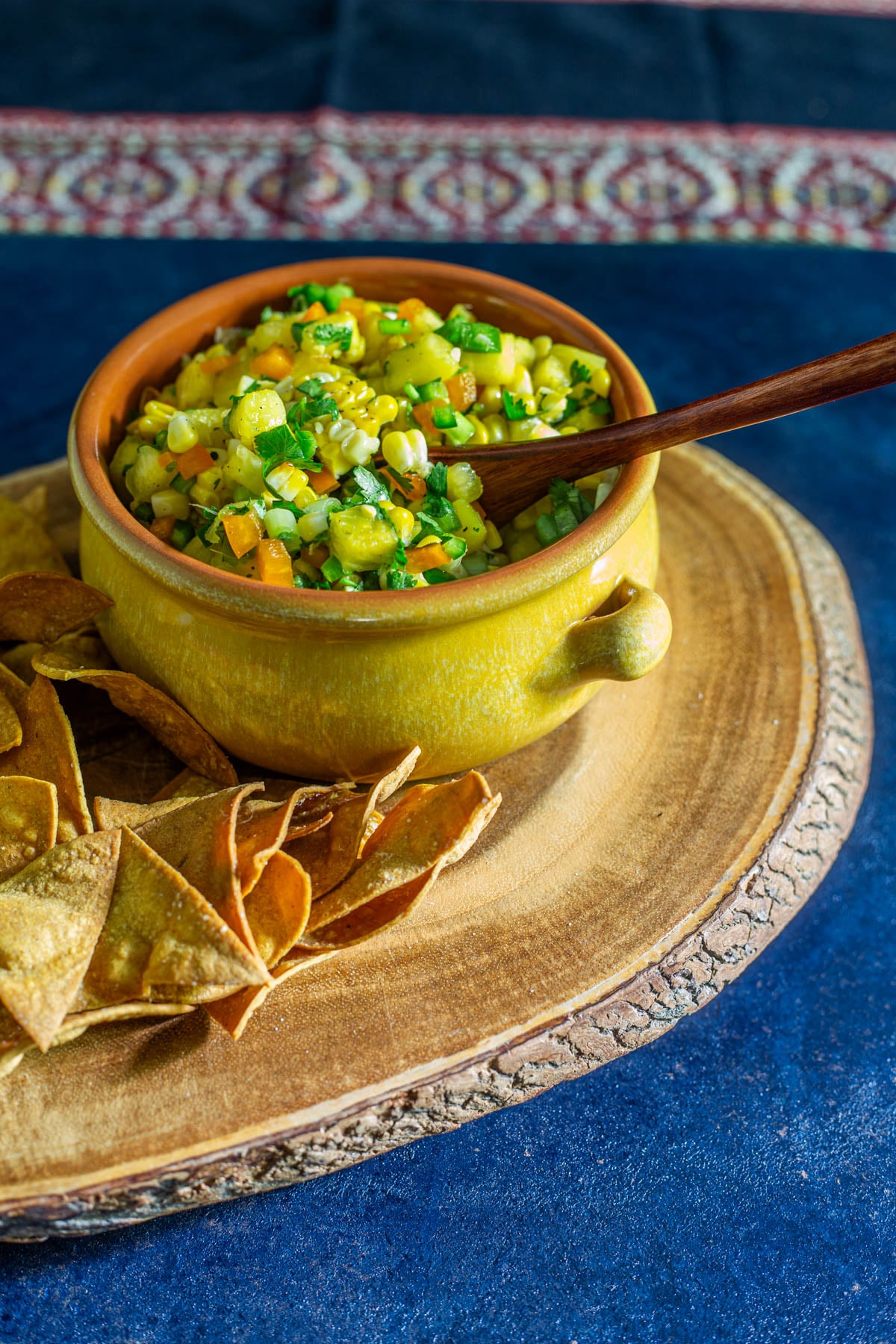 A yellow stoneware bowl with grilled corn and pineapple salsa on a live edge wood cutting board with tortilla chips.