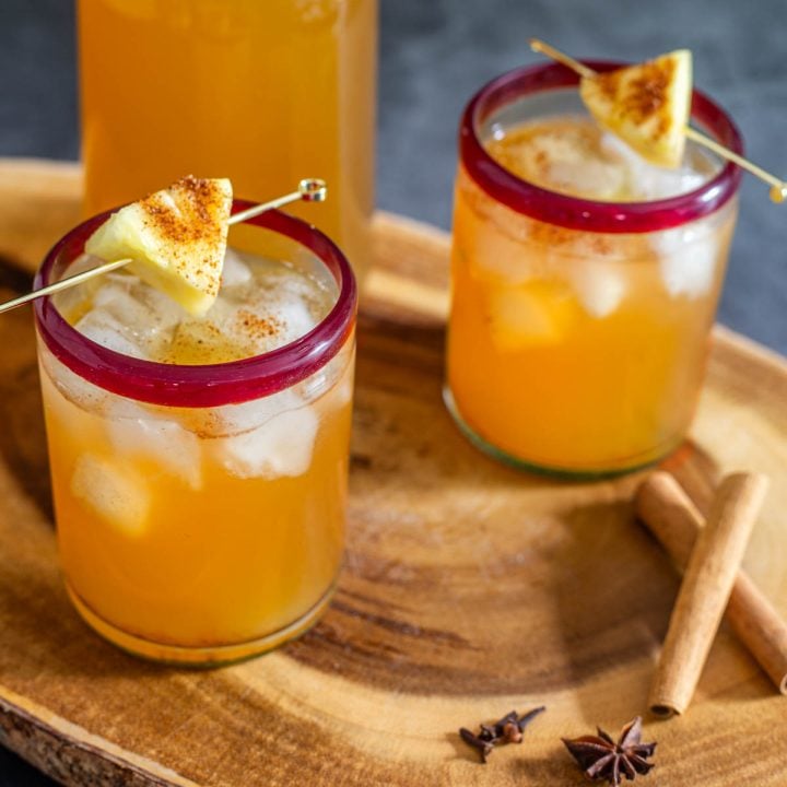 2 glasses of homemade tepache in red-rimmed glasses on a wood tray.