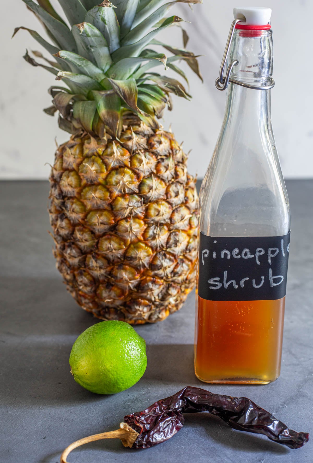 A swing-top glass bottle with 1 1/2 cups of Latin-inspired pineapple shrub, a pineapple, a lime, and dried red chile.