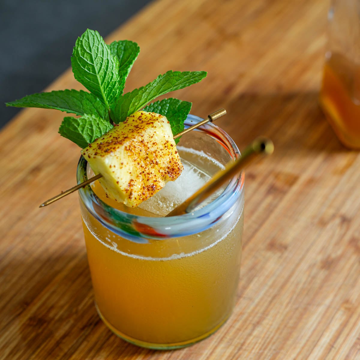 A close up of a pineapple shrub and soda garnished with a taj&iacute;n dusted pineapple chunk on a wood cutting board.
